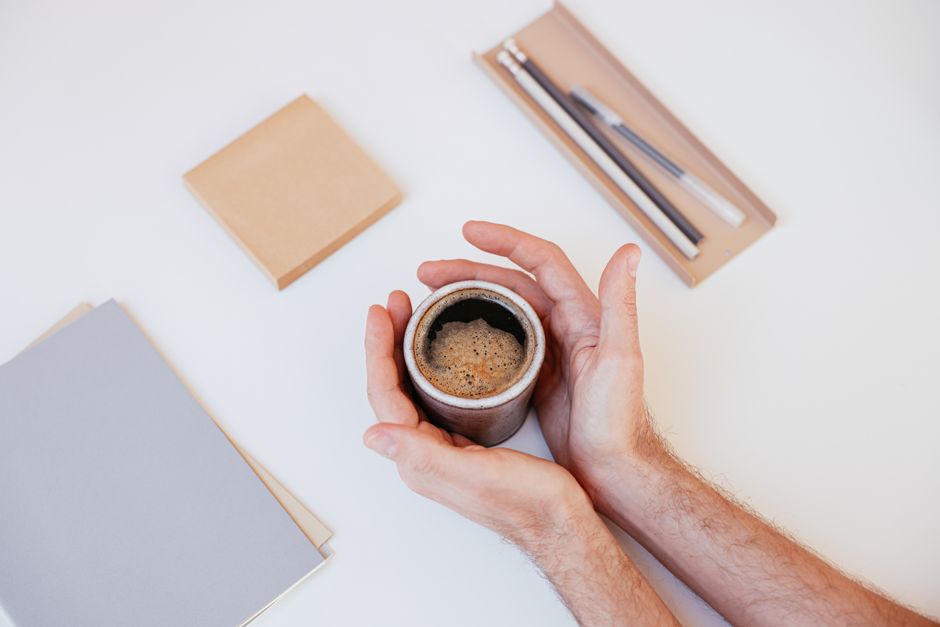 Hands holding a steaming cup of coffee beside writing materials on a white background.