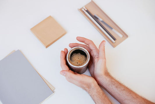 Hands holding a steaming cup of coffee beside writing materials on a white background.