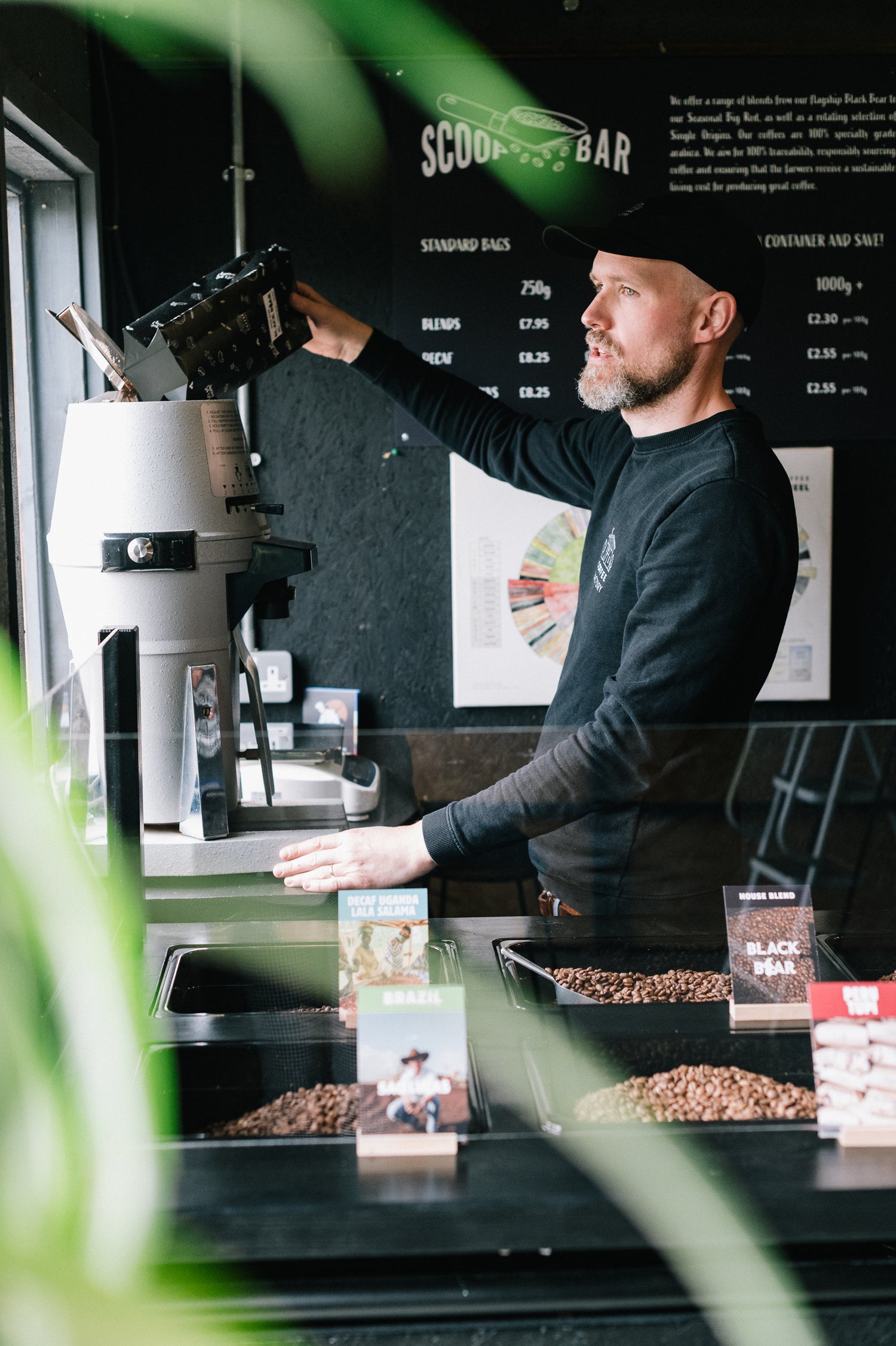 Danny Coffee Factory co founder at the Scoop Bar, pouring coffee from a grinder. Bean bins and product cards are visible on the counter, with blurred green leaves in the foreground.