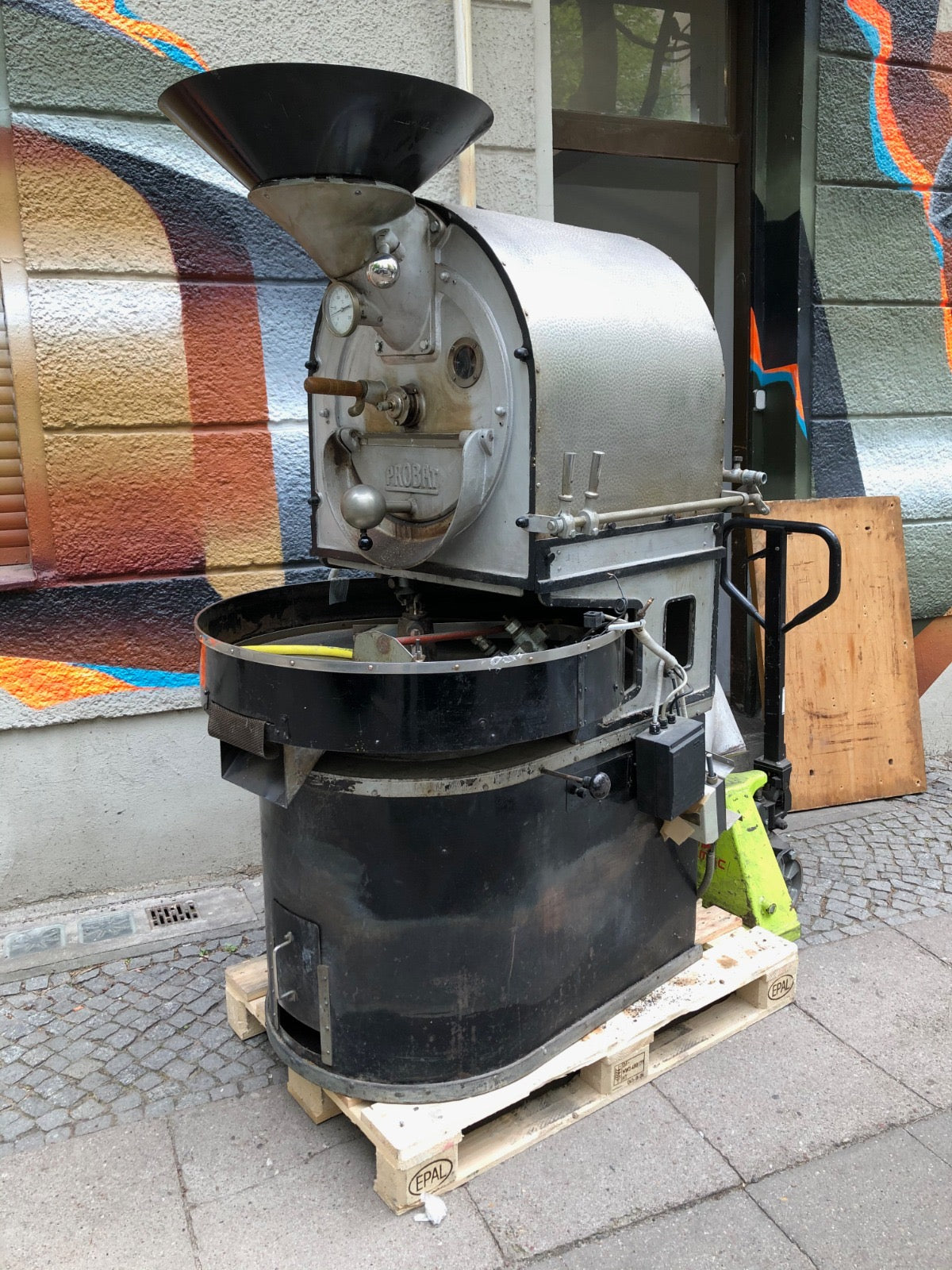 A large industrial coffee roaster machine on a wooden pallet, photographed outdoors on a paved street in front of a building with colorful graffiti.