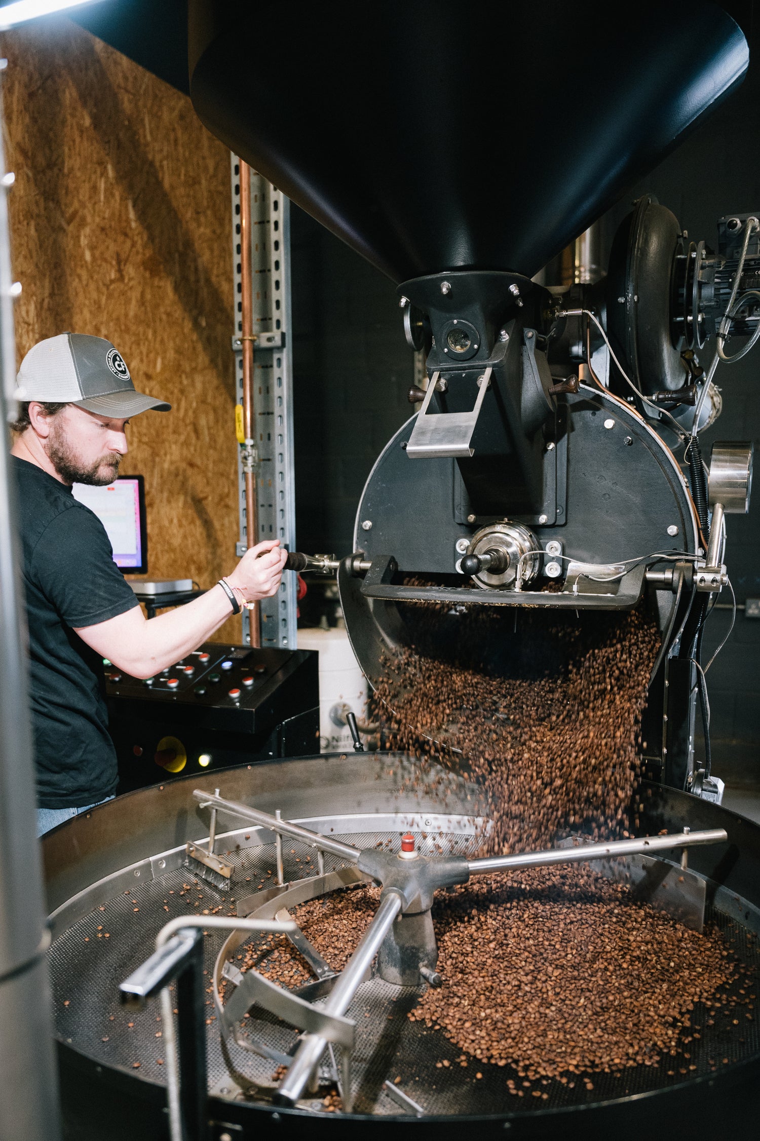 A man in a black t-shirt and baseball cap operates a large black coffee roasting machine, with roasted coffee beans pouring from the rotating drum into a collection tray below.